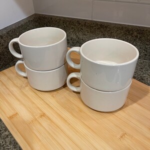 May include: Four white ceramic soup mugs stacked in pairs on a wooden cutting board. The mugs have a simple design and are perfect for serving soup or other hot beverages.