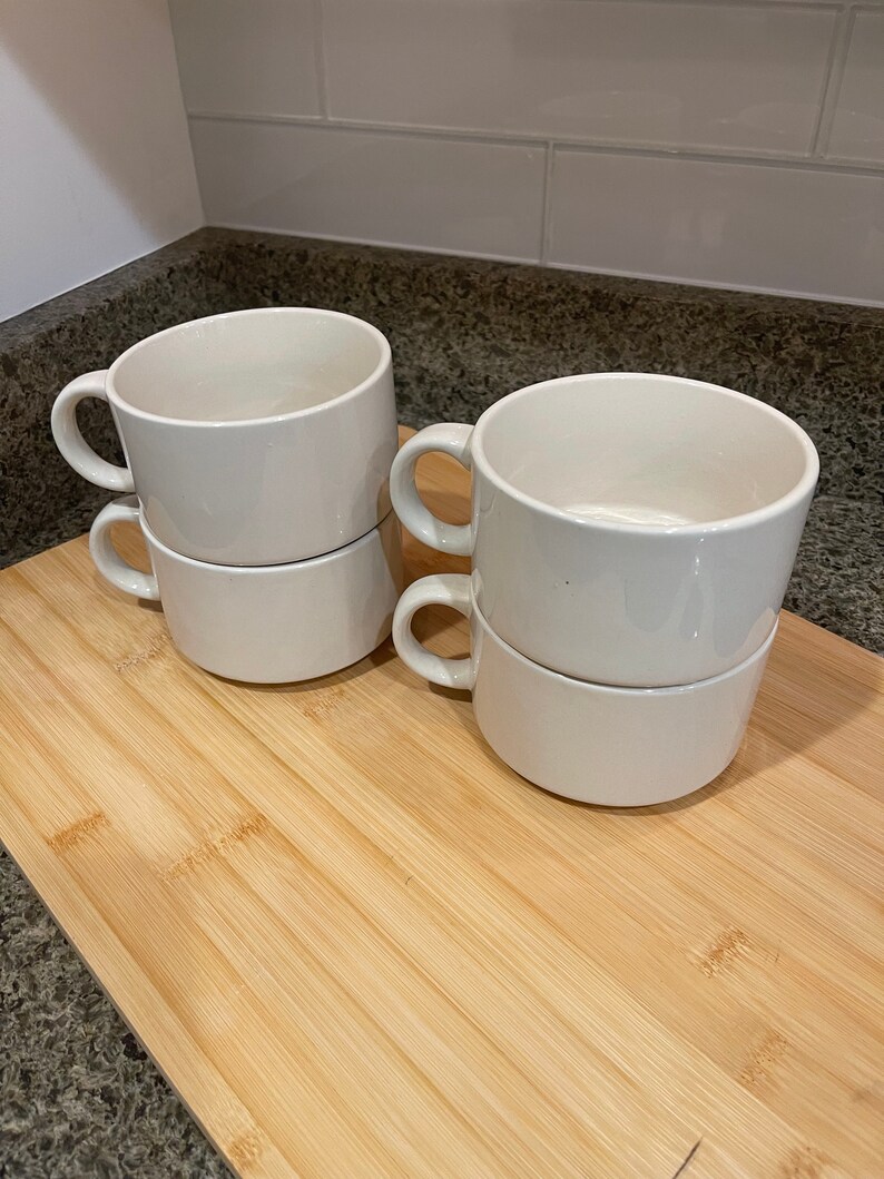 May include: Four white ceramic soup mugs stacked in pairs on a wooden cutting board. The mugs have a simple design and are perfect for serving soup or other hot beverages.