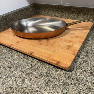May include: A copper-bottomed stainless steel frying pan with a long brass handle rests on a bamboo cutting board. The pan has a shiny interior and a rounded shape, ideal for cooking. The cutting board is rectangular.