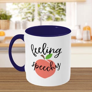 May include: White ceramic coffee mug with a blue rim. The mug has a peach graphic with a green leaf and the text "feeling peachy".