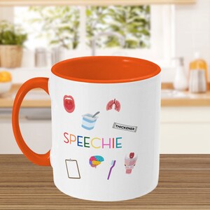 Puede incluir: Taza de café de cerámica blanca con borde y asa naranja. La taza tiene un diseño colorido con la palabra "SPEECHIE" y varios iconos relacionados con la logopedia, incluyendo una boca, pulmones, un cerebro, un portapapeles, un cepillo de dientes y un contenedor de yogur con la palabra "THICKENER" en una etiqueta.