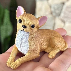 May include: A small, tan chihuahua figurine with white fur on its chest and pink ears. The figurine is lying down with its head tilted up.