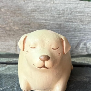 Könnte beinhalten: Beige Keramik-Hundefigur mit geschlossenen Augen und lächelndem Gesichtsausdruck. Der Hund hat eine braune Nase und Ohren. Die Figur ist ein dekoratives Objekt, möglicherweise für ein Regal oder einen Schreibtisch.