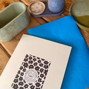 Peut inclure: Divers articles de décoration intérieure à motifs floraux, dont un plateau, une petite tasse, un dessous de verre et un récipient rectangulaire. Une feuille de papier bleu et une carte avec des autocollants de fleurs noirs et blancs sont également présents.