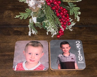 Puede incluir: Dos posavasos cuadrados de madera con fotos de niños pequeños. Los posavasos tienen un respaldo de corcho. El posavasos de la izquierda tiene una foto de un niño con cabello castaño que lleva un suéter rojo y blanco. El posavasos de la derecha tiene una foto de un niño con cabello castaño que lleva una camisa gris. El posavasos de la derecha también tiene el texto "2019 2020" impreso en él.