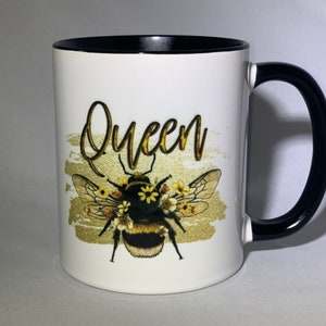 May include: White ceramic mug with a black handle and rim. The mug features a gold and black design of a bee with the word "Queen" in gold script. The bee is decorated with yellow flowers.