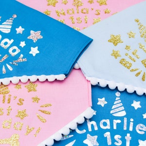 May include: Four dog bandanas with white pom pom trim. The bandanas are blue, pink, and light blue with gold glitter lettering. The text on the bandanas reads "Birthday Boy", "Cali's 1st Birthday", "Charlie's 1st", and "Lola's 1st".