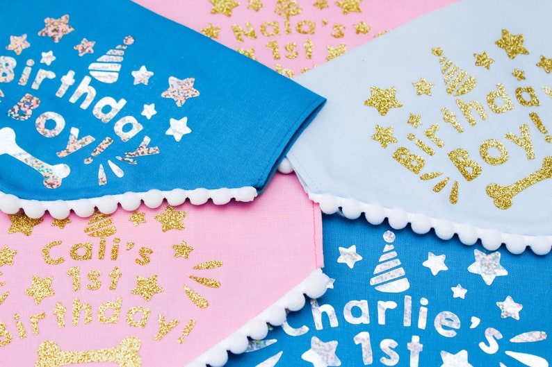 May include: Four dog bandanas with white pom pom trim. The bandanas are blue, pink, and light blue with gold glitter lettering. The text on the bandanas reads "Birthday Boy", "Cali's 1st Birthday", "Charlie's 1st", and "Lola's 1st".