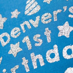 May include: A blue fabric background with silver glitter lettering that reads "Denver's 1st birthday". The lettering is surrounded by silver glitter stars.