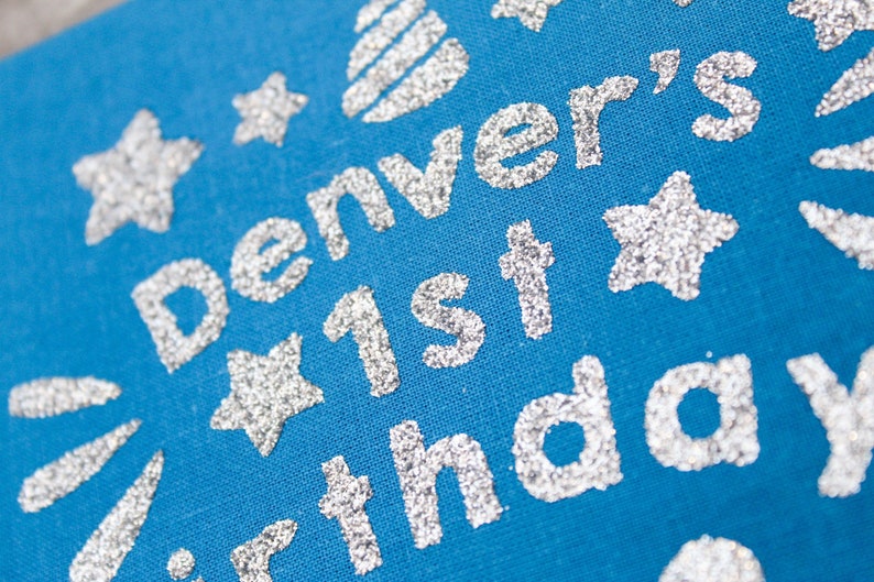 May include: A blue fabric background with silver glitter lettering that reads "Denver's 1st birthday". The lettering is surrounded by silver glitter stars.