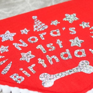 May include: Red dog bandana with white trim and silver glitter lettering that says "Nora's 1st Birthday!" with a bone and stars.