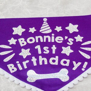May include: Purple dog bandana with white glitter lettering that says "Bonnie's 1st Birthday!" with a white glitter party hat and stars. The bandana has a white lace trim.