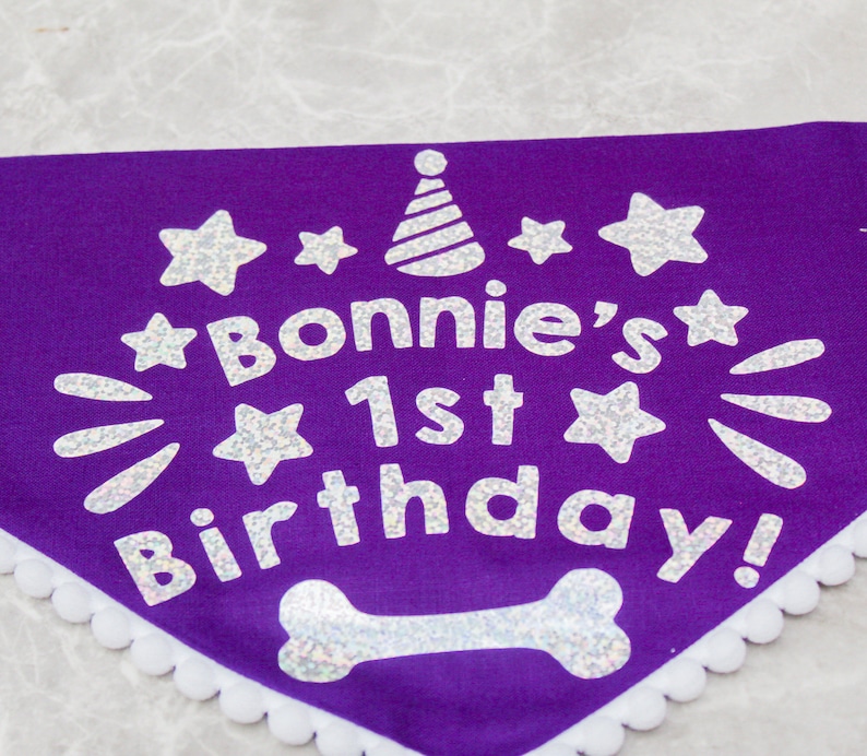 May include: Purple dog bandana with white glitter lettering that says "Bonnie's 1st Birthday!" with a white glitter party hat and stars. The bandana has a white lace trim.