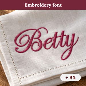 Peut inclure: Une serviette en lin de couleur crème avec le prénom Betty brodé dans une police cursive rose foncé. La serviette présente un détail de bordure à ourlet ajouré. Le texte Embroidery font et + BX sont visibles sur l'image.