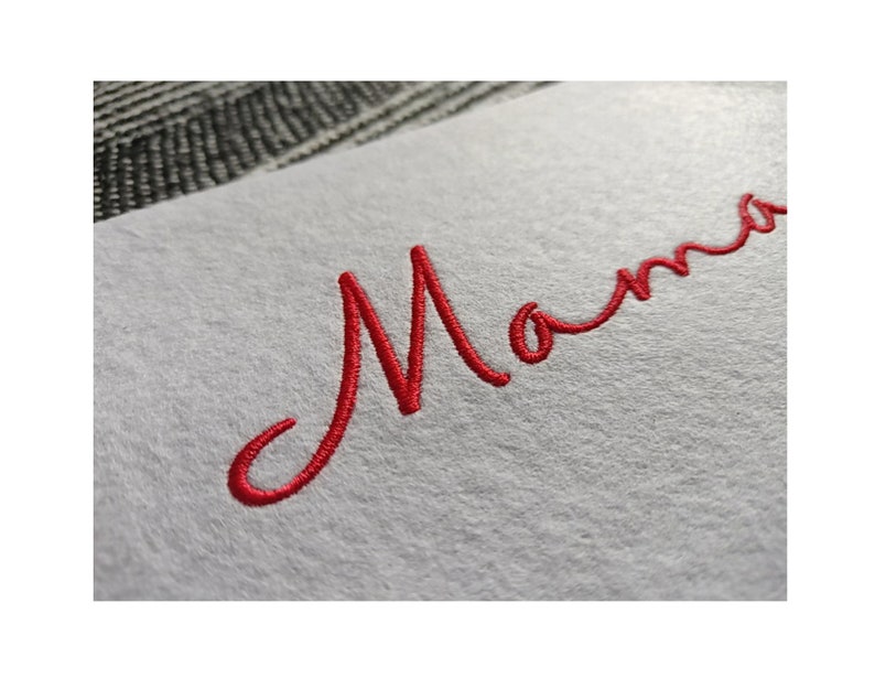 K&ouml;nnte beinhalten: Rote gestickte kursive Schrift, die "Mama" auf einem wei&szlig;en Stoffhintergrund liest.