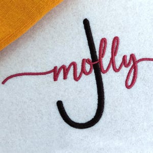 May include: A white fabric surface featuring machine embroidery with the name molly stitched in a cursive red font. A large, vertical black letter J is embroidered over the name. A corner of textured mustard yellow fabric is visible at the top.