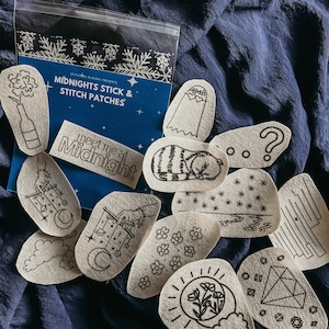 May include: A set of 12 felt patches with various designs, including a ghost, a cat, a question mark, a flower, a moon, a diamond, and a hand holding a string of stars and moons. The patches are packaged in a clear plastic bag with a blue label that reads "Midnights Stick & Stitch Patches" and "Meet me at Midnight".