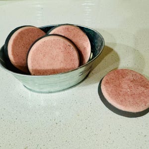 May include: Four round pink soap bars with black edges in a silver metal bowl, and one similar soap bar on a white countertop.