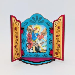 May include: A wooden diorama featuring a religious image of an angel with wings, holding a scale and sword. The diorama has a turquoise frame with floral cutouts, red and gold side panels with rose designs, and a pink base.