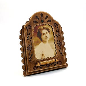 May include: Wooden picture frame with a sepia-toned portrait of a woman. The frame is brown with a scalloped edge and floral vine detailing. The text "GRANDMA ANN" is on a banner at the bottom. A small green gem is at the top.