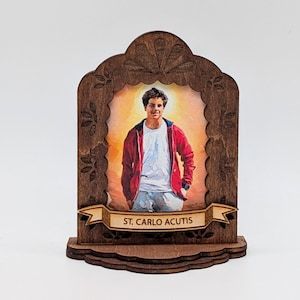 May include: Wooden religious icon featuring a portrait of St. Carlo Acutis. The image shows a young man in a red jacket and white shirt, set against a warm, glowing background. The icon is framed with ornate, carved wooden details and includes a banner that reads "ST. CARLO ACUTIS."