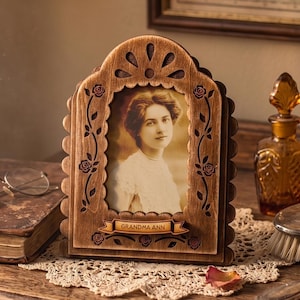 May include: An arched wooden picture frame with a dark brown finish, featuring laser-cut rose motifs along the sides and a decorative top. A banner at the bottom displays the text GRANDMA ANN. The frame holds a sepia-toned portrait of a person.