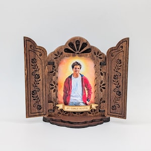 May include: A wooden triptych featuring a portrait of St. Carlo Acutis. The central image is framed by a decorative wooden structure with floral and leaf patterns. The portrait shows a young man in a red jacket and white shirt. The text "ST. CARLO ACUTIS" is on a banner.