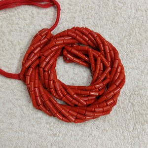 Op de afbeelding: Een verzameling levendige rode koraalkralen, geregen op een rood koord. De kralen zijn cilindrisch en gelijkmatig van formaat, waardoor een cirkelvormige opstelling ontstaat. Het koraal heeft een gepolijste, glanzende afwerking en het koord is bovenaan zichtbaar.