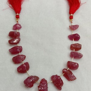 May include: A strand of raw, deep pink ruby gemstones strung on a thin gold-colored wire. The strand features irregular, faceted stone shapes and is finished at both ends with bright red decorative tassels attached by small gold-toned metal caps.
