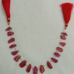 Puede incluir: Un hilo de piedras preciosas en bruto de forma irregular y color rojo rosado, ensartadas en un alambre fino de color dorado. El hilo termina en ambos extremos con una borla decorativa de color rojo brillante con un pequeño casquillo dorado.