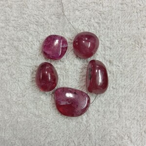 May include: A strand of five polished, irregular, deep pinkish-red gemstone beads. Each bead features a smooth, glossy surface with natural internal inclusions and variations in colour intensity, strung together on a thin, metallic silver-coloured wire.
