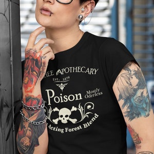 Puede incluir: Camiseta negra con un dise&ntilde;o gr&aacute;fico blanco que dice "Thee Apothecary Est. 1874 Poison Mostly Odorless Acting Forest Blend" con una calavera y huesos cruzados.