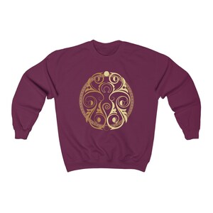 May include: A maroon sweatshirt with a gold circular design featuring intricate floral patterns and a repeating motif. The design is centered on the front of the sweatshirt.