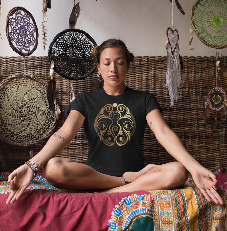 May include: A woman wearing a black t-shirt with a gold mandala design. The mandala features a circular pattern with intricate details and a crescent moon shape.