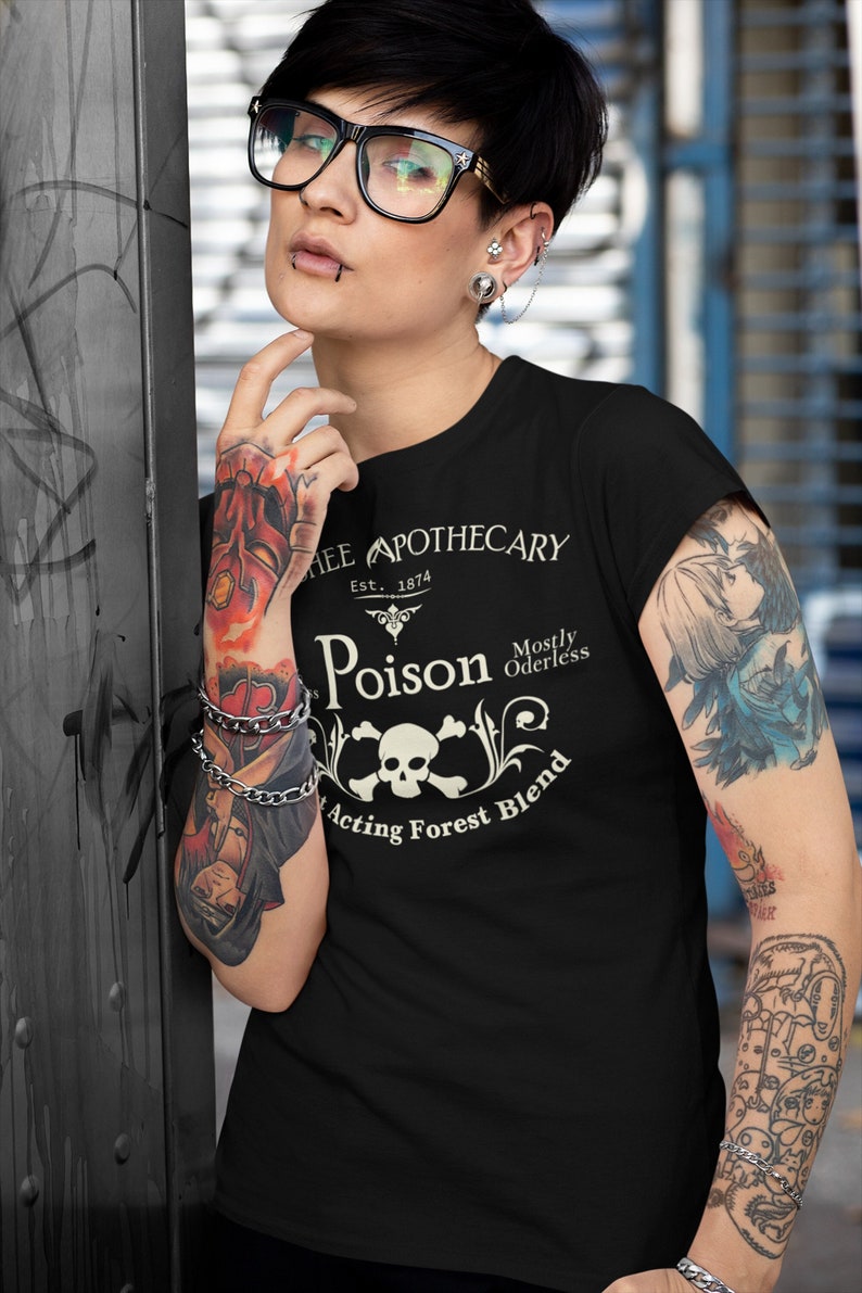 Puede incluir: Camiseta negra con un dise&ntilde;o gr&aacute;fico blanco que dice "Thee Apothecary Est. 1874 Poison Mostly Odorless Acting Forest Blend" con una calavera y huesos cruzados.