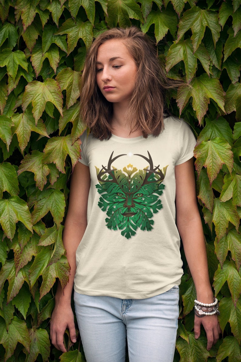 May include: A white t-shirt with a green and brown leafy design of a face with antlers. The design is reminiscent of the Green Man, a mythical figure associated with nature.