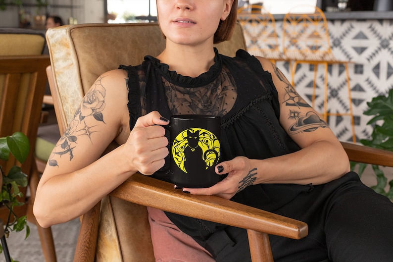 May include: A black ceramic mug with a yellow silhouette of a cat sitting in a circle of floral designs.