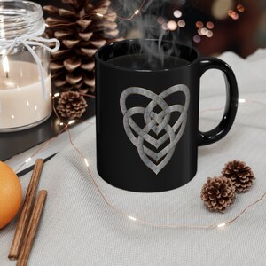 Op de afbeelding: Zwarte keramische mok met een zilveren Keltisch knoop hartontwerp. De mok is gevuld met koffie en er stijgt stoom op uit de bovenkant.