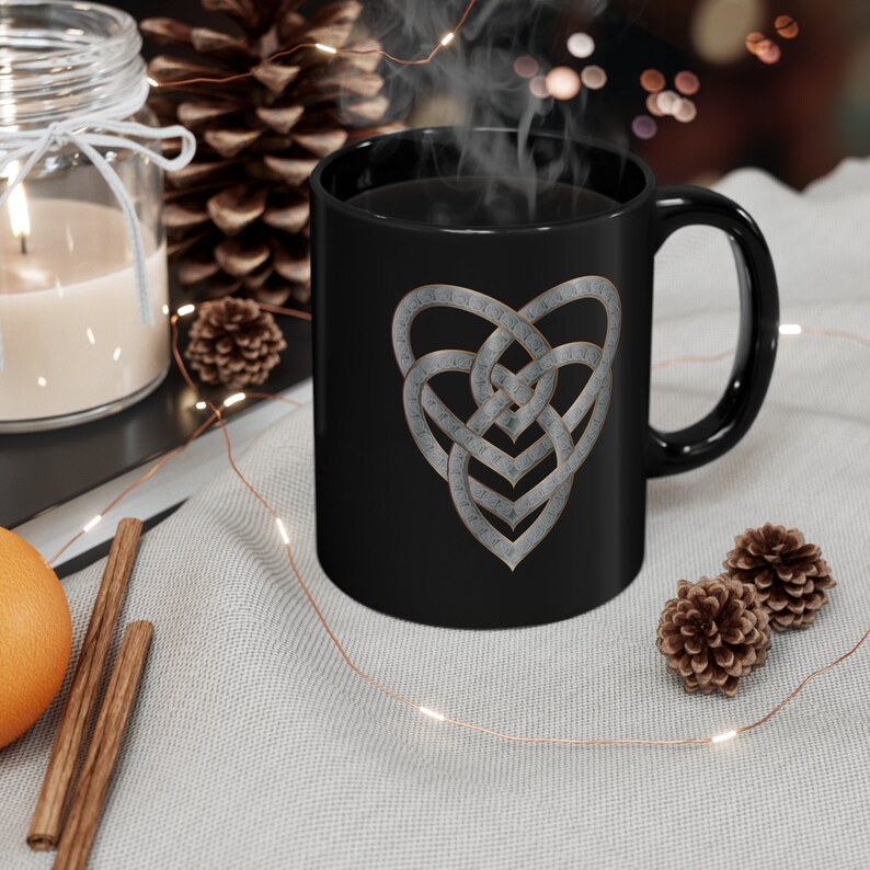 Op de afbeelding: Zwarte keramische mok met een zilveren Keltisch knoop hartontwerp. De mok is gevuld met koffie en er stijgt stoom op uit de bovenkant.