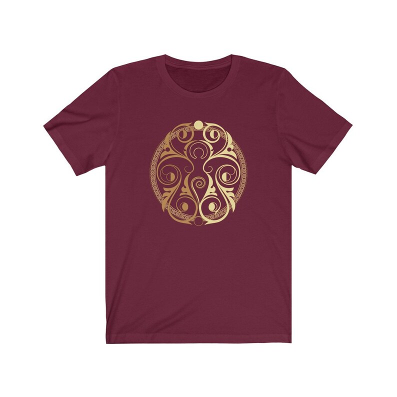 May include: A maroon t-shirt with a gold circular design featuring a stylized floral pattern and crescent moons.