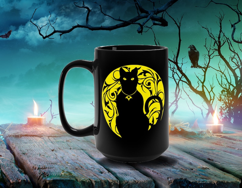 May include: Black coffee mug featuring a black cat silhouette on a yellow moon with floral designs. The mug is on a wooden surface with a candle in the background.