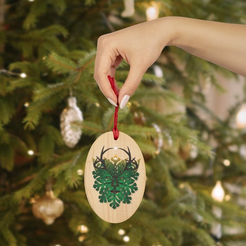 Peut inclure: Une d&eacute;coration de No&euml;l ovale en bois avec un motif vert et or d'un Homme Vert avec des bois. L'ornement est suspendu &agrave; une ficelle rouge.