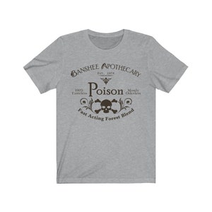 Puede incluir: Una camiseta gris con un dise&ntilde;o gr&aacute;fico negro que dice "Banshee Apothecary Est. 1874 Poison 100% Tasteless Mostly Oderless Fast Acting Forest Blend". El gr&aacute;fico tambi&eacute;n incluye una calavera y huesos cruzados.