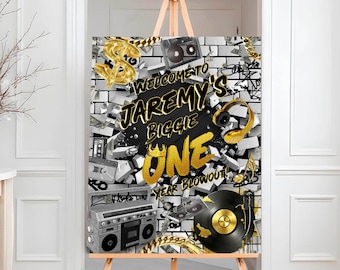 May include: A black and white birthday party sign with gold accents. The sign features a brick wall background with a gold dollar sign, a cassette tape, a record player, and headphones. The text on the sign reads "Welcome to Jaremy's Biggie One Year Blowout!"
