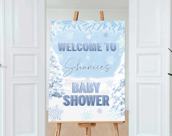 May include: A light blue baby shower sign on a wooden easel. The sign reads "Welcome to Shanice's Baby Shower" with a winter theme, featuring snowflakes and snowy trees. The easel is made of light brown wood.