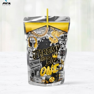 May include: A silver pouch with a yellow straw sticking out of the top. The pouch has a black and white design with gold accents. The design includes a dollar sign, headphones, a record player, a boombox, and the text "Jaremy the Big One".