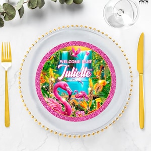 Puede incluir: Un plato decorativo con un borde de purpurina rosa y un diseño tropical con flamencos, cascadas y exuberante vegetación. El plato tiene el texto "WELCOME BABY Juliette". Un tenedor y un cuchillo dorados están a un lado.