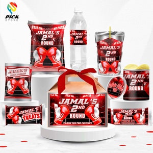 May include: A boxing-themed party favor set with red and black labels featuring the name "Jamal's 2nd Round". The set includes a box, a bag of chips, a juice box, a water bottle, a chocolate bar, and a can of potato chips.