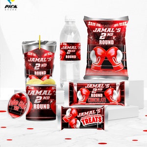 May include: A set of party favors for a boxing themed birthday party. The favors include a water bottle, a juice box, a bag of chips, a chocolate bar, and a box of marshmallow treats. All of the items have a red and black boxing theme with the text "Jamal's 2nd Round" on them.