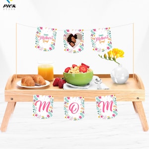 Puede incluir: Una bandeja de desayuno de madera con una pancarta floral que dice "¡Feliz Día de la Madre!" y "MAMÁ". La pancarta está atada con una cuerda y tiene una foto de una madre y un niño en un marco en forma de corazón. La bandeja también tiene un tazón de cereal, un vaso de jugo de naranja y dos croissants.